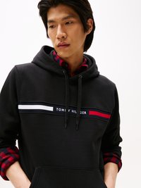 Felpa nera con cappuccio, decorata con strisce orizzontali bianche e rosse, con il logo "TOMMY HILFIGER", indossata sopra una camicia a quadri rossa e nera.