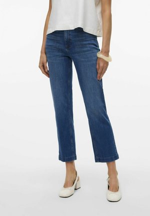 Blaue, verkürzte Jeans im geraden Schnitt mit leicht ausgefranstem Saum und klassischem Fünf-Taschen-Design. Getragen mit weißen High Heels.