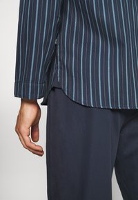 Camicia a righe navy con linee verticali azzurro chiaro, in tessuto di cotone, con orlo curvo e maniche lunghe, abbinata a pantaloni scuri e lisci.