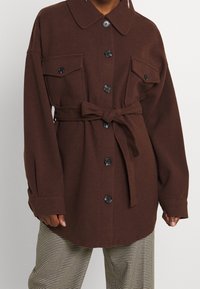 Hnědá košile na knoflíky z měkké látky s límečkem, dvěma náprsnými kapsami a zavazovacím páskem v pase. Černé knoflíky dodávají kontrast.