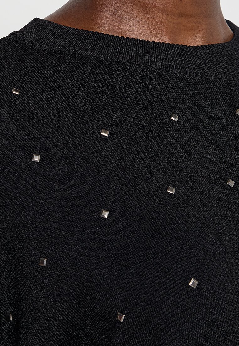 Nahaufnahme eines schwarzen Strickstoffs mit kleinen silbernen quadratischen Nieten, die verstreut sind und den Ausschnitt sowie den oberen Brustbereich einer Person zeigen.