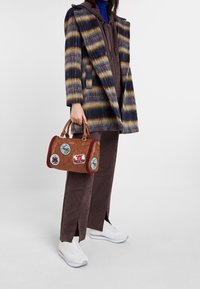 Bruine leren handtas met rode accenten en patches, textuurafwerking, voorzien van korte handvatten en een gestructureerde vorm. Gestreepte jas en neutrale broek.