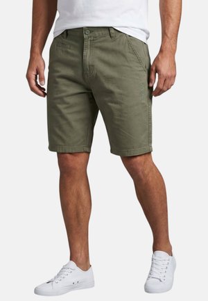 Homme portant un short olive de longueur genou, un T-shirt blanc et des baskets blanches, debout les jambes légèrement écartées.