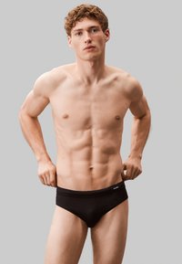 Bañadores negros de corte ajustado con una textura suave y una cinturilla elástica, que presentan un pequeño logotipo de la marca en la parte frontal. El modelo está de pie con las manos en la cintura.