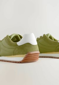 Grønne ruskind- og stof sneakers med hvide hælaccenter, der har en tekstureret brun gummisål og kontrasterende hvide og gule detaljer.