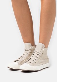 High-top beige canvas sneakers with white rubber toe cap and sole, featuring prominent laces and stitching details.