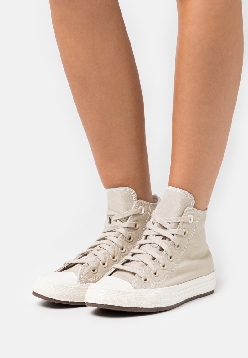 High-top beige canvas sneakers with white rubber toe cap and sole, featuring prominent laces and stitching details.