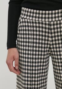 Pantalon à carreaux noir et crème, avec une texture de tissu lisse et une ceinture ajustée. Main reposant sur le côté.