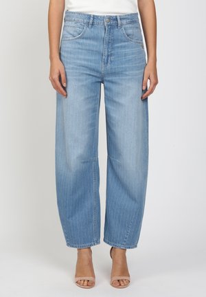 Frau trägt hellblaue, hoch taillierte Jeans mit weitem Bein und dezenten vertikalen Streifen sowie nudefarbene offene Absatzschuhe.