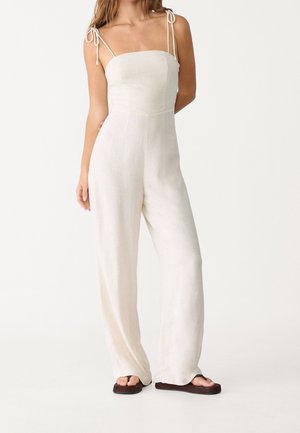 Combinaison en lin couleur crème avec des bretelles fines et ajustables, corsage ajusté et pantalons à jambes larges. Texture lisse, design minimal.