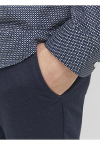 Granatowa koszula z białym geometrycznym wzorem, z mankietem zapinanym na guziki oraz widoczną kieszenią na ciemnoniebieskich spodniach. Gładka tekstura tkaniny.