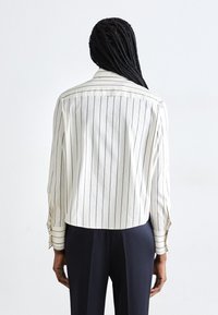 Witte gestreepte shirt met blauwe verticale lijnen, met een afgeronde zoom, lange mouwen en knoopsluiting bij de manchetten. De stof lijkt glad en licht van gewicht.