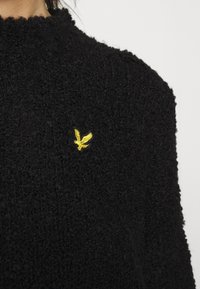 Maglione nero con texture e collo alto, caratterizzato da un piccolo logo di un'aquila gialla ricamata sul petto. Tessuto morbido e peloso.