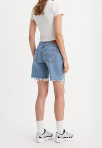 Pantalones cortos de mezclilla con dobladillo deshilachado, color azul claro, diseño de cinco bolsillos y una etiqueta de cuero marrón en la parte posterior. Llevados con una camiseta blanca ajustada.