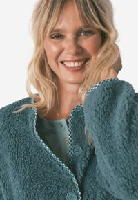 Türkisfarbener, flauschiger Cardigan mit strukturiertem Finish, runden Knöpfen und einem gemusterten Besatz am Rundhalsausschnitt. Unterlage aus Baumwollmischung sichtbar.