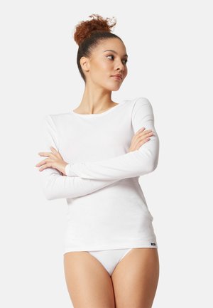 Valkea pitkähiha-paita, valmistettu pehmeästä kankaasta, jossa pyöreä kaula-aukko. Malli poseeraa käsivarret ristissä, yllään vastaavat valkoiset alushousut.