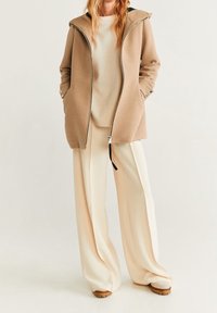 Femme portant un manteau zippé beige, un pull crème, un pantalon large crème et des chaussures beiges, se tenant devant un fond uni.