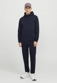 Navy Hoodie mit Reißverschluss vorne und zwei Seitentaschen, kombiniert mit passenden navy Jogginghosen. Weiße Sportschuhe und eine helllila Kappe vervollständigen das Outfit.