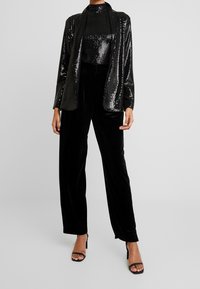 Černý flitrový blazer přes černý rolák, spárovaný s volnými černými sametovými kalhotami a černými vysokými podpatky, vytvářející elegantní siluetu.