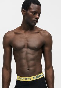 Μυώδης άνδρας χωρίς μπλούζα που κοιτάζει προς τα κάτω, φορώντας μαύρα εσώρουχα με κίτρινη ζώνη που γράφει «BJÖRN SWEDEN» σε απλό γκρι φόντο.
