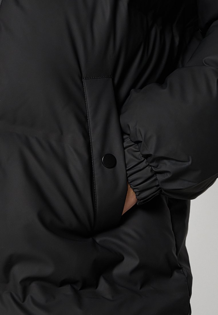 Doudoune noire avec une texture lisse et brillante. Présente un poignet élastiqué et une poche latérale sécurisée avec une fermeture à pression.