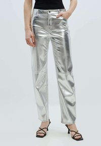 Zilveren metallic broek met een gladde, glanzende textuur, voorzien van een rechtlijnig ontwerp en zijzakken. Gepaard met zwarte sandaaltjes met bandjes.