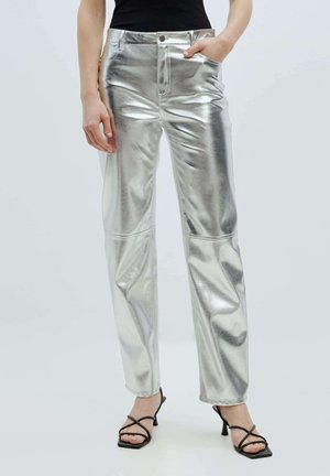Zilveren metallic broek met een gladde, glanzende textuur, voorzien van een rechtlijnig ontwerp en zijzakken. Gepaard met zwarte sandaaltjes met bandjes.