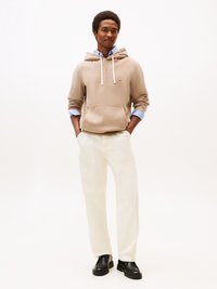 Felpa beige con cappuccio e lacci, indossata sopra una camicia azzurra chiara, abbinata a pantaloni bianchi e scarpe nere, in piedi su uno sfondo neutro.