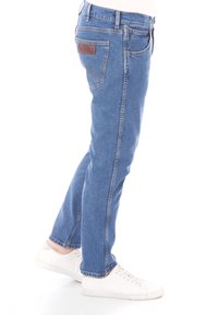 Jeans de mezclilla azul con un corte recto, cinco bolsillos y un parche de cuero marrón en la parte trasera de la cintura. Combinados con zapatillas blancas.