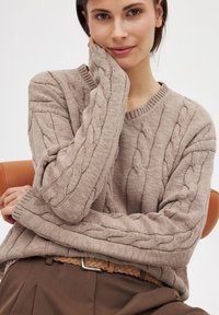 Bežinis megztinis su V formos iškirpte, su tekstūrinėmis pynėmis ir ilgomis rankovėmis, derinamas su aukšto liemens rudi kelnių ir pynimo diržu.