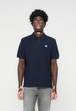 Polo de color azul marino con cuello, de manga corta y un pequeño parche con logo; combinado con vaqueros ligeros grises.