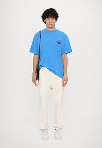 Blå bomullströja med korta ärmar och en bröstgrafik, kombinerad med krämfärgade sweatpants och vita sneakers. Svart axelremsväska.