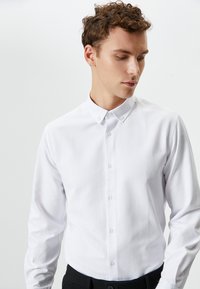 Camisa blanca de botones con pequeños puntos texturizados, de mangas largas y con cuello. Llevada con pantalones oscuros, que presentan un diseño de corte estándar.