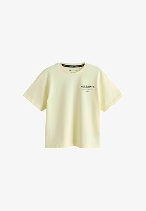 Niña joven de pie con una camiseta color amarillo pálido de AllSaints, pantalones cortos de estampado de leopardo, calcetines blancos y zapatillas negras sobre un fondo gris.