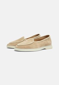 Mocasines de ante beige con punta redondeada, detalles de costura sutiles en la parte superior y una suela de goma blanca para mayor amortiguación y agarre.