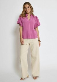 Blusa rosa a maniche corte con scollatura a V e dettagli raccolti, abbinata a pantaloni beige a gamba larga e tacchi. Tessuto morbido, tessuto leggero.