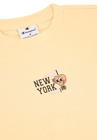Jasnożółta bluza z okrągłym dekoltem i krótkimi rękawami. Zawiera postać z kreskówki oraz napis "NEW YORK SINCE 1919" w czarnym kolorze.