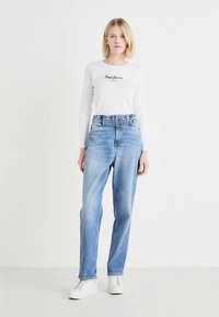 Bela majica z dolgimi rokavi s črnim napisom, združena s svetlo modrimi kavbojkami z visokim pasom in belimi supergami. Model stoji pred nevtralno ozadje.