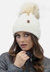 Witte gebreide beanie met een grote nep-bont pompon, voorzien van een gestructureerd patroon en een klein bruin merklabel aan de voorkant.
