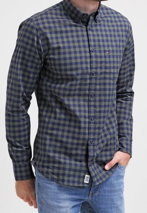 Mann trägt ein langärmliges, marineblau und olivgrün kariertes Hemd mit Knöpfen, blaue Jeans, Hand in der Tasche, vor einem einfarbigen Hintergrund.