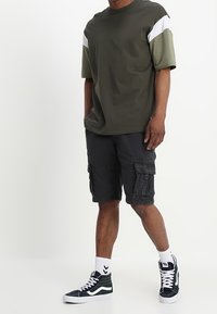 Camiseta de algodón verde oscuro con acentos en blanco y oliva, combinada con pantalones cortos cargo negros, calcetines blancos y zapatillas verdes y blancas.