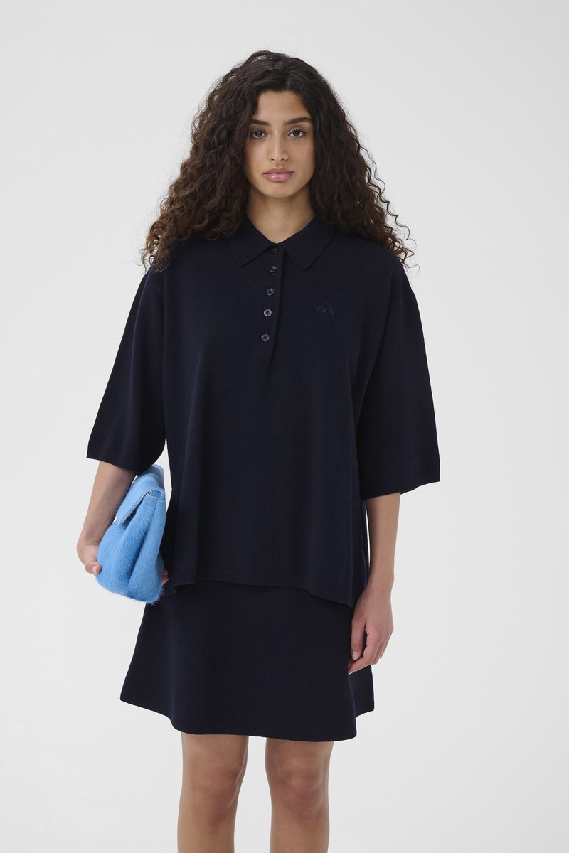 Top polo tricotat bleumarin cu mâneci scurte, cu o deschidere cu cinci nasturi, și fustă asortată. Modelul ține o geantă texturată albastru deschis.