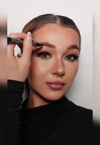Une femme à la peau lisse et claire applique un crayon à sourcils foncé. Elle porte un haut ajusté à manches longues noir, avec de longs cheveux brillants.