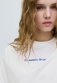 Biała bawełniana koszulka o swobodnym kroju. Na przodzie niebieskie haftowane napisy "Off-season. On pu." Prosty, minimalistyczny design.