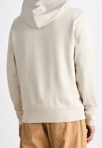Felpa beige con cappuccio regolabile, texture liscia e orlo a costine. Abbinata a pantaloni marrone chiaro, vista da dietro.