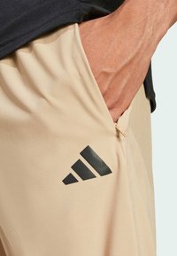 Beige sportbroek met een gladde textuur, voorzien van een zwart Adidas-logo aan de zijkant en een ritszak op de heup.