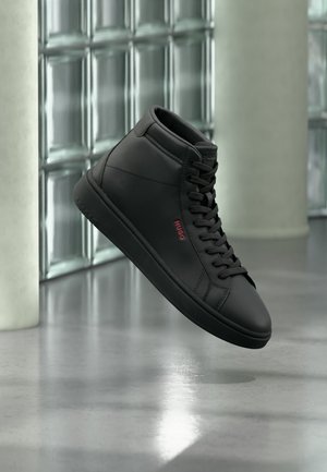 Schwarzer High-Top-Sneaker mit Schnürsenkeln und rotem "Hugo"-Logo, dargestellt vor einem Hintergrund aus Glasbausteinen und Betonboden.