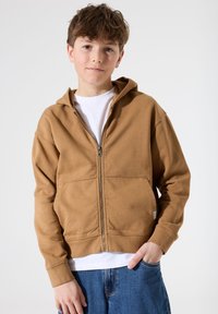 Sudadera con cremallera marrón hecha de tela suave, con un bolsillo tipo canguro y un corte holgado, llevada sobre una camiseta blanca y combinada con jeans azules.