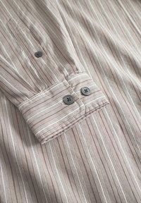 Cămașă bej cu dungi subțiri albe, cu o textură moale. Manșete cu nasturi cu două butoane închise la culoare și o guler bine definit.