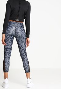 Woman in black long-sleeve top and patterned gray leggings with mesh panels, wearing white Puma sneakers, standing against white background.